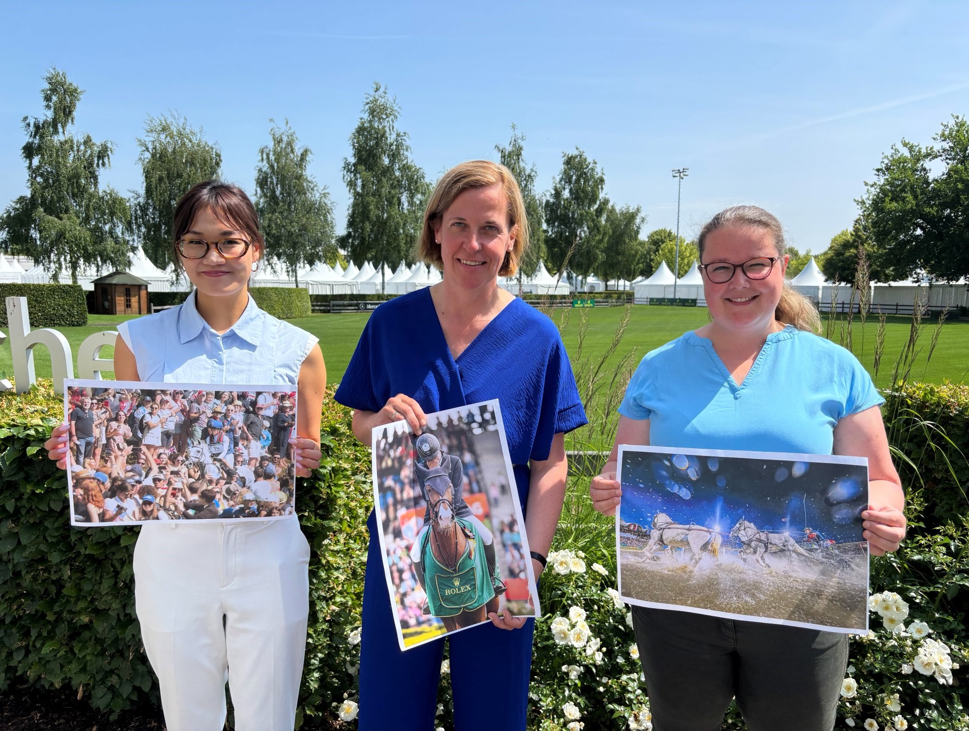 Drei Personen stehen nebeneinander im Freien vor blühenden weißen Rosen und halten jeweils ein großes Farbfoto vor sich: Die linke Person präsentiert ein Bild mit einer jubelnden Menschenmenge, die mittlere Person hält ein Foto mit einem Reiter auf einem Pferd, die rechte Person zeigt ein Bild mit einer Pferdeszene im Wasser. Im Hintergrund sind grüne Wiesen, Bäume und eine Reihe weißer Pagodenzelte zu sehen.