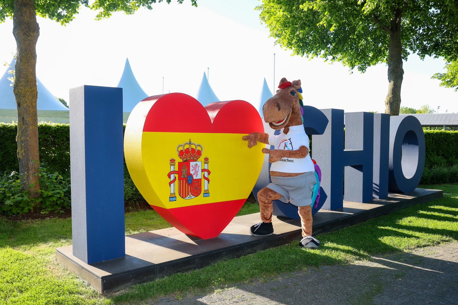 Das CHIO Aachen Maskottchen, ein fröhliches Pferd in weißem T-Shirt und grauer Hose, steht vor einer großen Skulptur mit der Aufschrift „I Love CHIO“. Das Herz in der Mitte ist in den Farben der spanischen Flagge gestaltet und trägt das spanische Wappen. Im Hintergrund sind Bäume, Büsche und weiße Zeltspitzen zu sehen.
