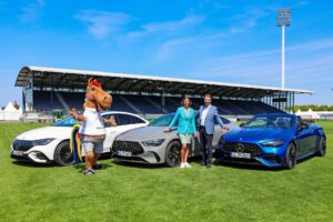 Drei moderne Mercedes-Benz Fahrzeuge stehen auf dem Rasen eines Stadions. Neben den Autos posieren ein Maskottchen in Pferdekostüm sowie zwei elegant gekleidete Personen. Im Hintergrund ist eine überdachte Tribüne mit dem Mercedes-Benz Logo zu sehen, darüber ein klarer blauer Himmel.