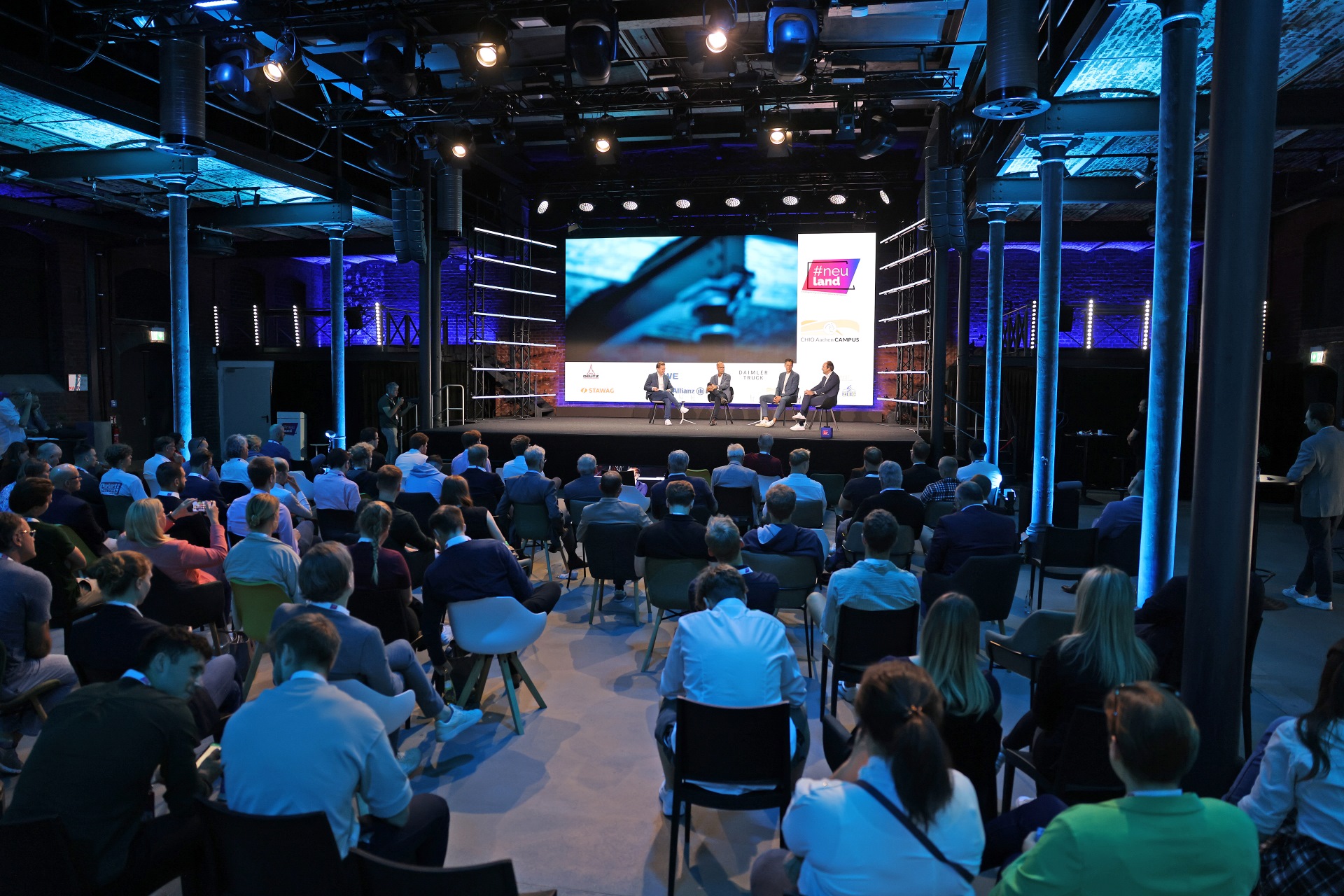 Ein Kongress in einer modern umgestalteten Industriehalle mit atmosphärischer blauer Beleuchtung. Auf der Bühne findet eine Podiumsdiskussion mit vier Personen statt, die auf Stühlen sitzen. Zahlreiche Zuschauer in Business-Kleidung sitzen im Auditorium und verfolgen aufmerksam die Diskussion.