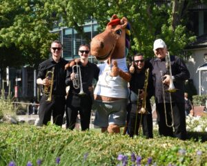 A group of four musicians dressed in black stand in the greenery with trumpets and trombones next to a large, cheerful horse mascot wearing a white T-shirt labelled #chioaachen.