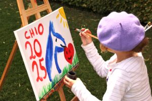 Ein Kind mit einer lila Baskenmütze malt im Freien an einer Staffelei ein buntes Bild. Auf der Leinwand sind ein blaues Pferd, ein rotes Herz mit Gesicht, eine gelbe Sonne und die roten Buchstaben und Zahlen „CHIO 2025“ zu sehen. Das Kind hält einen Pinsel in der rechten Hand und trägt ein gestreiftes Langarmshirt.