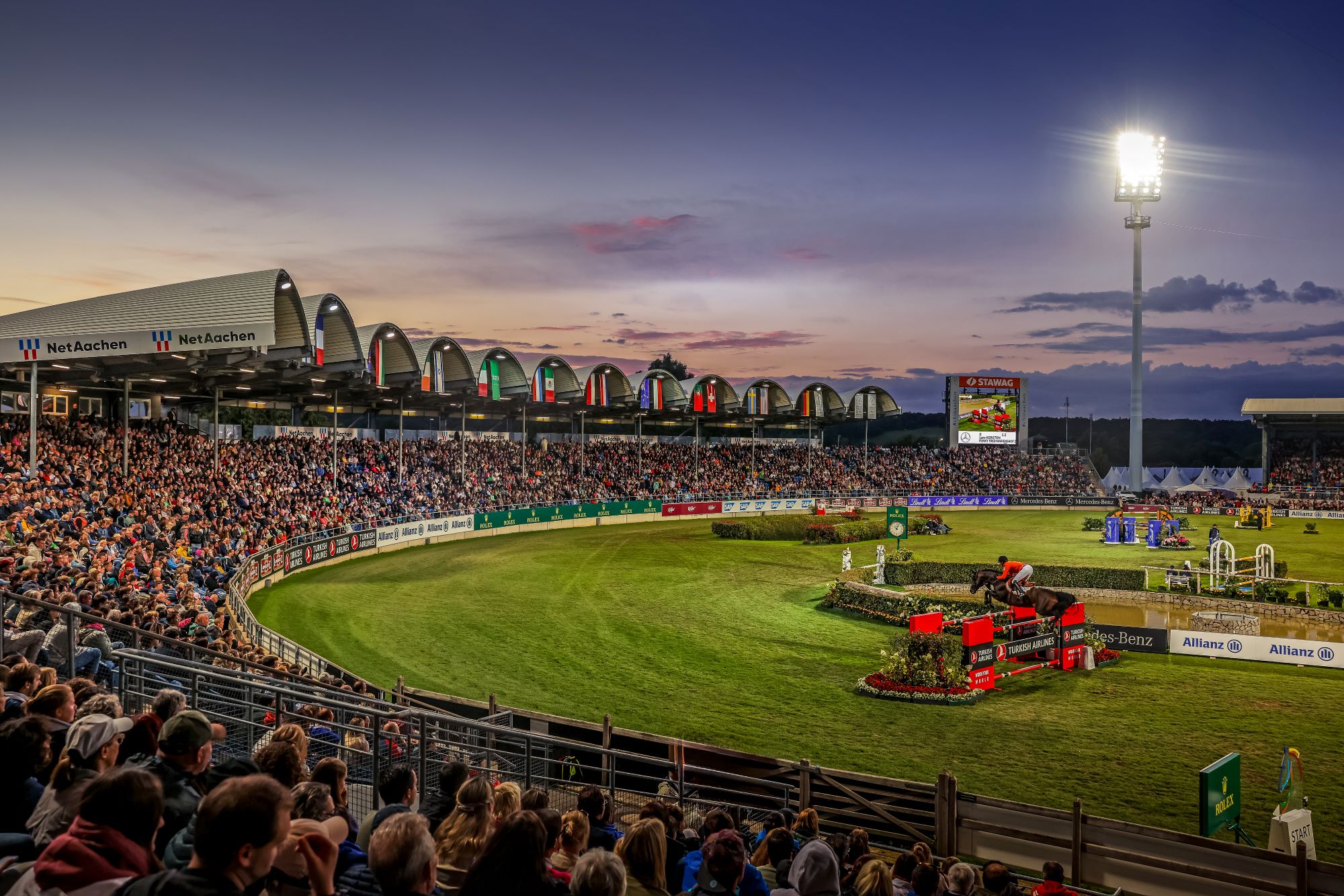 Großes Reitstadion bei Sonnenuntergang, voll besetzte Tribünen und beleuchtetes Springturnier. Im Vordergrund springt ein Reiter mit seinem Pferd über ein Hindernis, während das Publikum das Geschehen aufmerksam verfolgt.