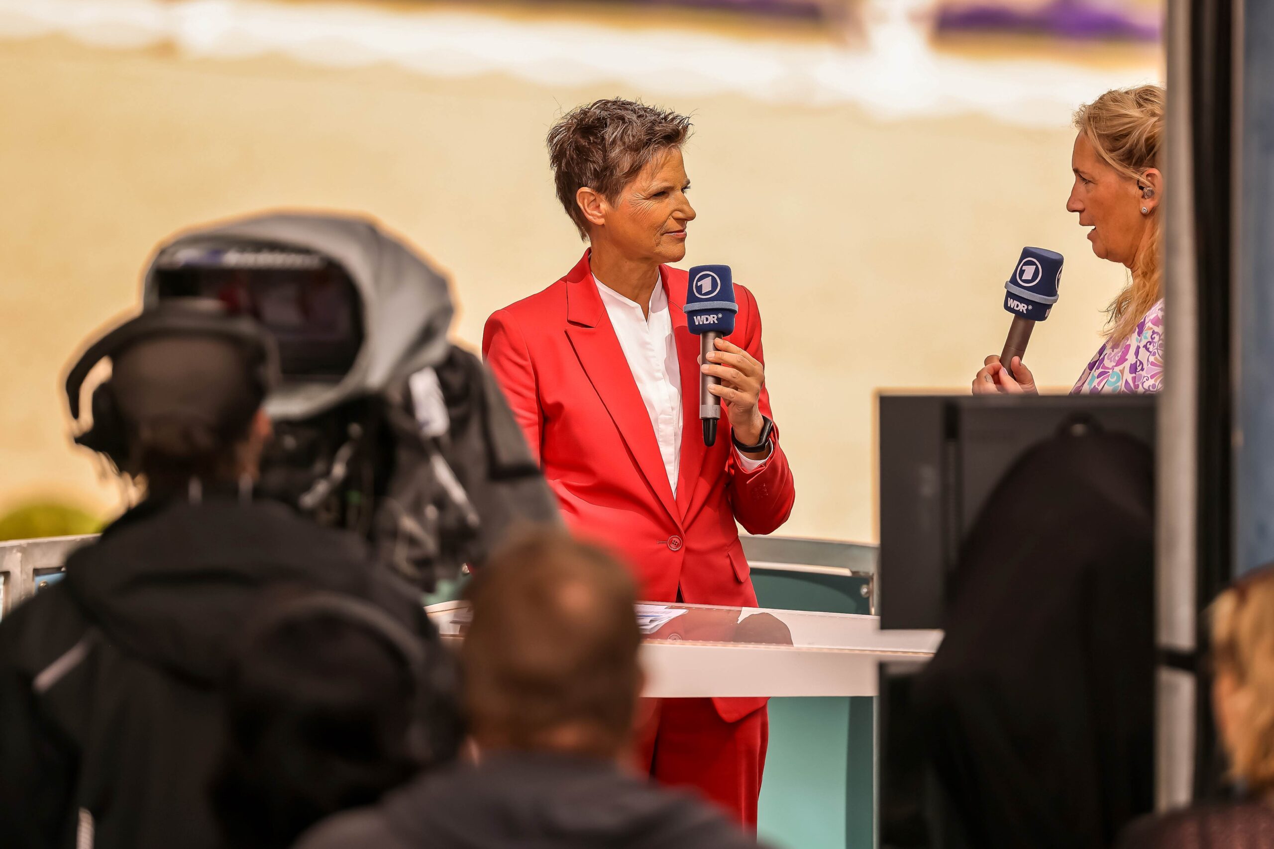 Zwei Frauen stehen in einem Fernsehstudio an einem Tisch und halten Mikrofone mit dem Logo von WDR und ARD. Eine Person trägt einen roten Anzug, die andere ein gemustertes Oberteil. Im Vordergrund ist ein Kamerateam zu sehen, im Hintergrund lässt sich unscharf das Dressurstadion erahnen.