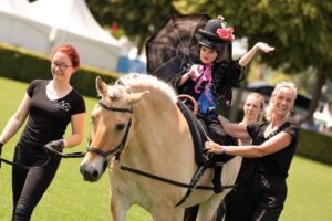A young girl performing therapeutic riding in a costume with a black hat, lace, colourful ribbons and a black parasol sits on a Haflinger. Two adults dressed in black lead the horse and support the child from the side. White tents and trees can be seen in the background.