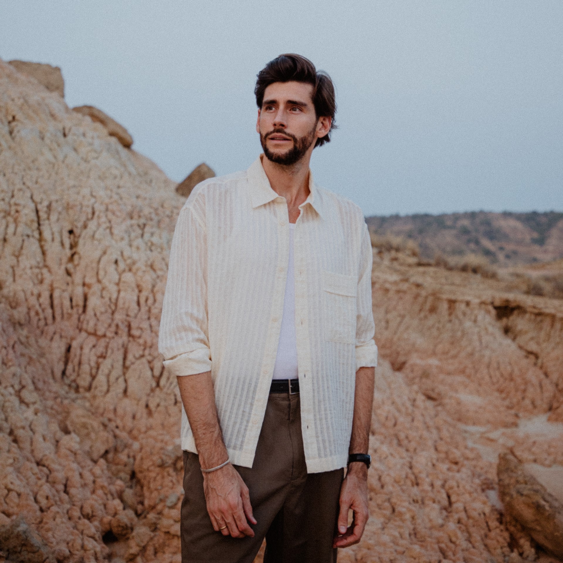 Alvaro Soler, bekleidet mit einem offenen, cremefarbenen gestreiften Hemd über einem weißen T-Shirt und braunen Hosen, steht zwischen rauen Felsformationen vor einem blassen Himmel und blickt nachdenklich in die Ferne.