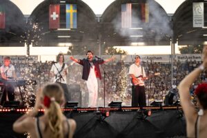 Auf einer großen Bühne in einem Stadion steht eine Band und spielt live vor einem begeisterten Publikum. Im Vordergrund sind Tänzerinnen mit Blumen im Haar. Der Sänger in weißer Hose und dunkler Jacke steht mit ausgebreiteten Armen im Zentrum, umgeben von Bandmitgliedern an Keyboard, Bass und Gitarre. Funken sprühen auf der Bühne, im Hintergrund sind Fahnen verschiedener Nationen und die vollbesetzten Tribünen zu sehen.