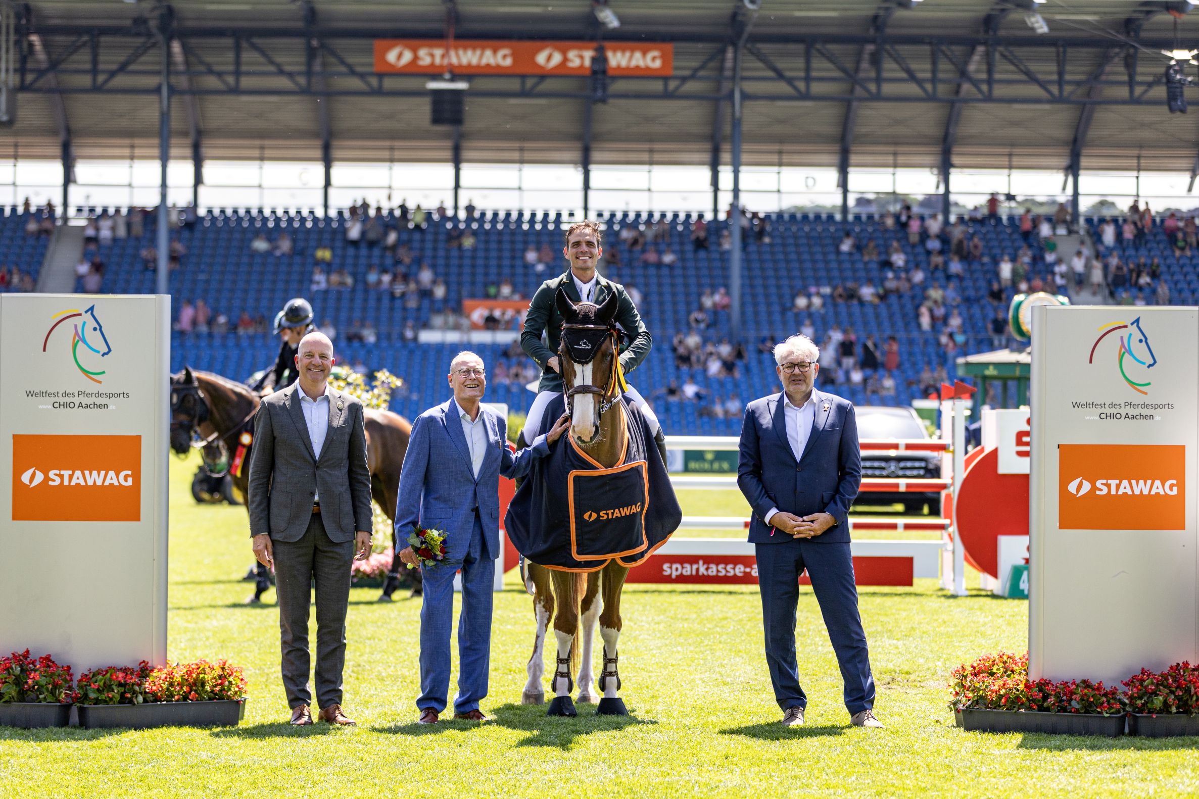 The photo shows a rider in a green jacket on a brown horse with a white blaze and four white legs. The horse is wearing a dark blue winner's rug with orange edging and the STAWAG logo. To the left and right of the horse are three men in suits, one holding a bouquet of flowers. In the background, blue stands with spectators, obstacles and large signs with the CHIO Aachen logo and STAWAG lettering can be seen. The scene takes place on the sunny grass arena.