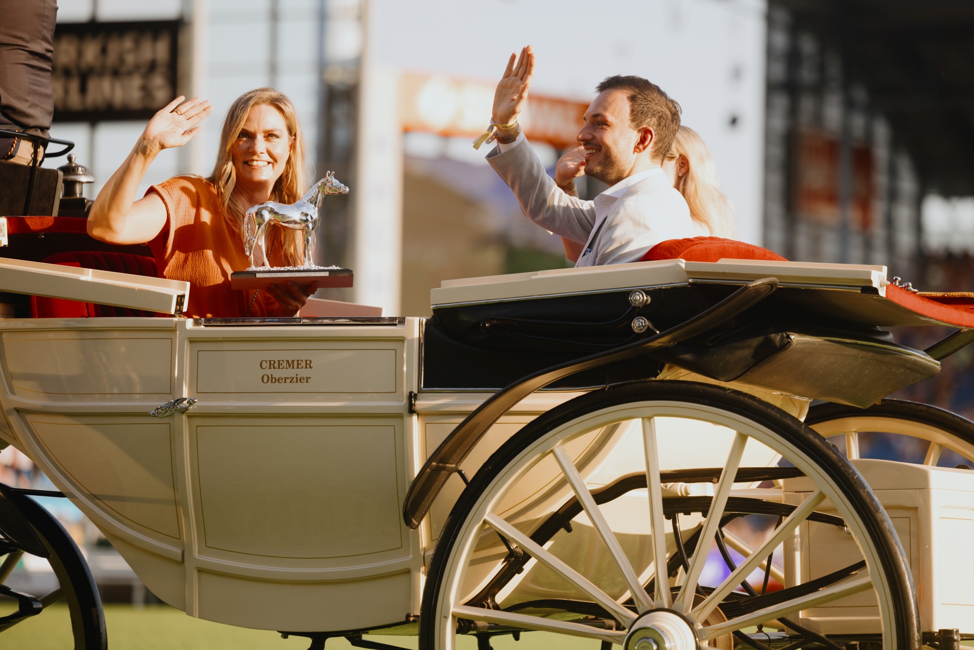 People sitting in a cream-colored horse-drawn carriage wave to the crowd. One person is holding a silver horse trophy. The carriage has the name "CREMER Oberzier" written on the side, and the scene takes place in a stadium with sunlight illuminating the group.