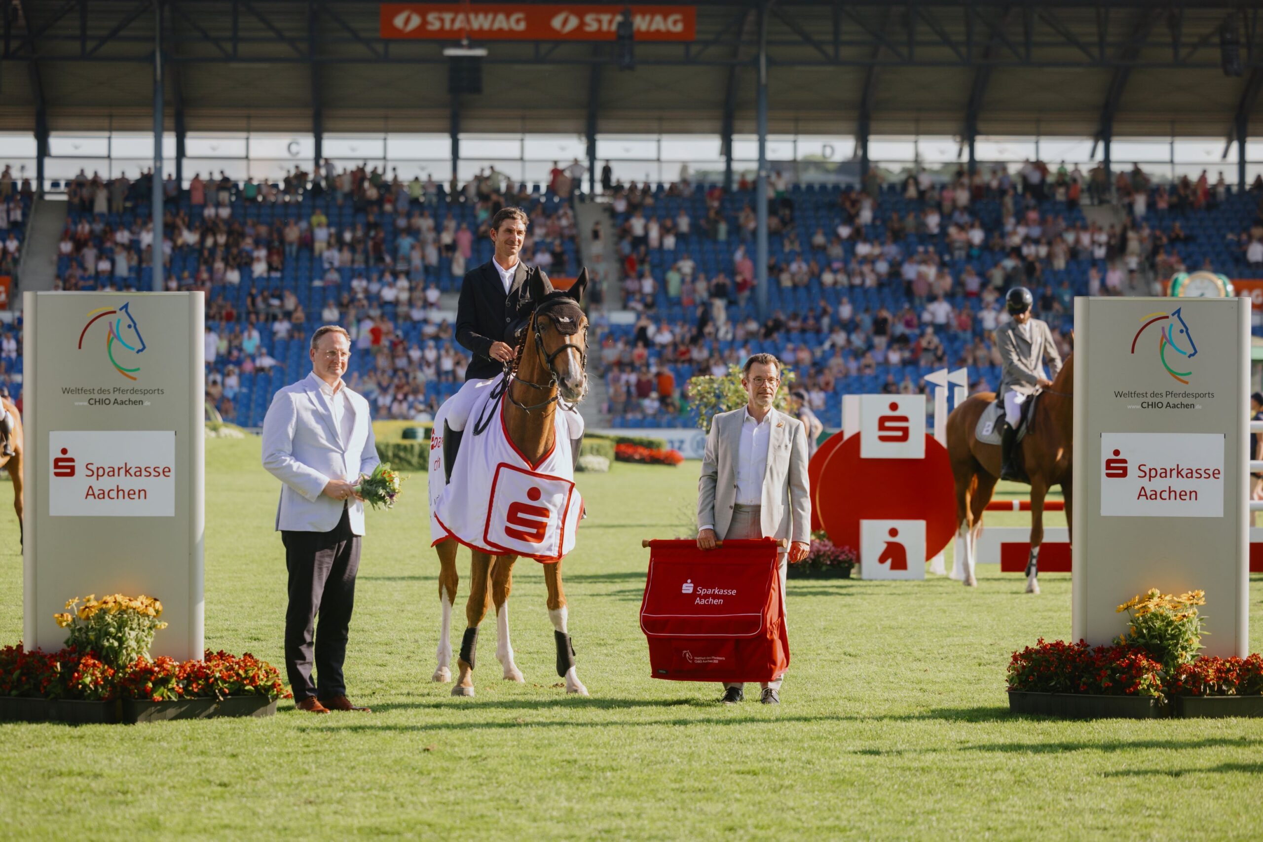 A rider sits on a horse wearing a winner's blanket with the Sparkasse logo. To the left and right, two men in suits stand, one holding a bouquet of flowers, the other holding a red bag labeled "Sparkasse Aachen". In the background, there are more riders and a large crowd of spectators in the stands. The scene takes place on the green grass of a stadium, framed by advertising boards for Sparkasse and CHIO Aachen.