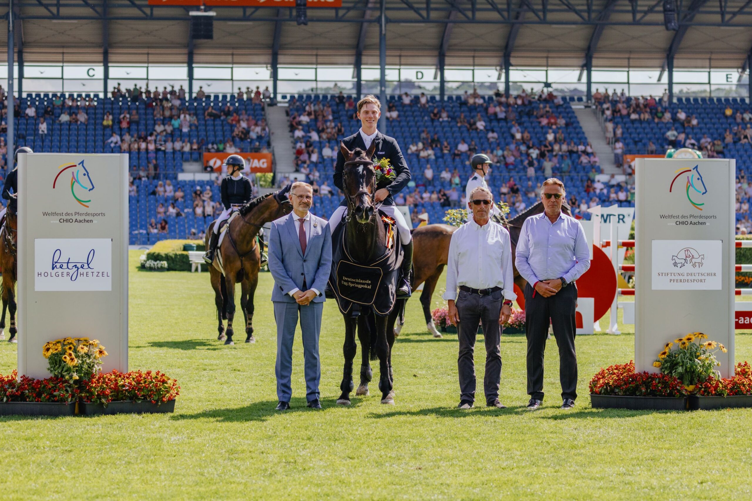 A rider in a dark jacket sits on a dark brown horse with a black winner's blanket bearing the words ‘Deutschlands U25 Springpokal’ in gold lettering. The rider is holding a bouquet of flowers in his hand. To the left and right of the horse are three men, one in a blue suit and the others in light blue shirts. In the background are more riders, spectator stands with lots of people and two large signs with the CHIO Aachen logo and sponsor logos. In front of the signs are floral decorations and colourful flowers.