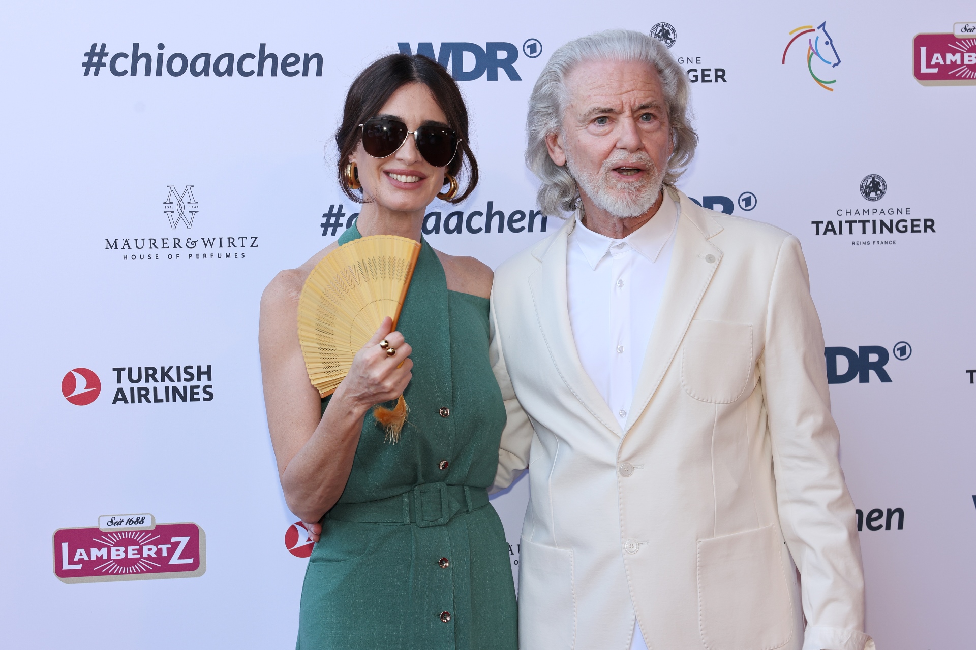 A woman in a green, off-the-shoulder dress holds a yellow fan in her hand and stands next to a man in a white suit. Both are posing together in front of a sponsor wall at the CHIO Aachen.
