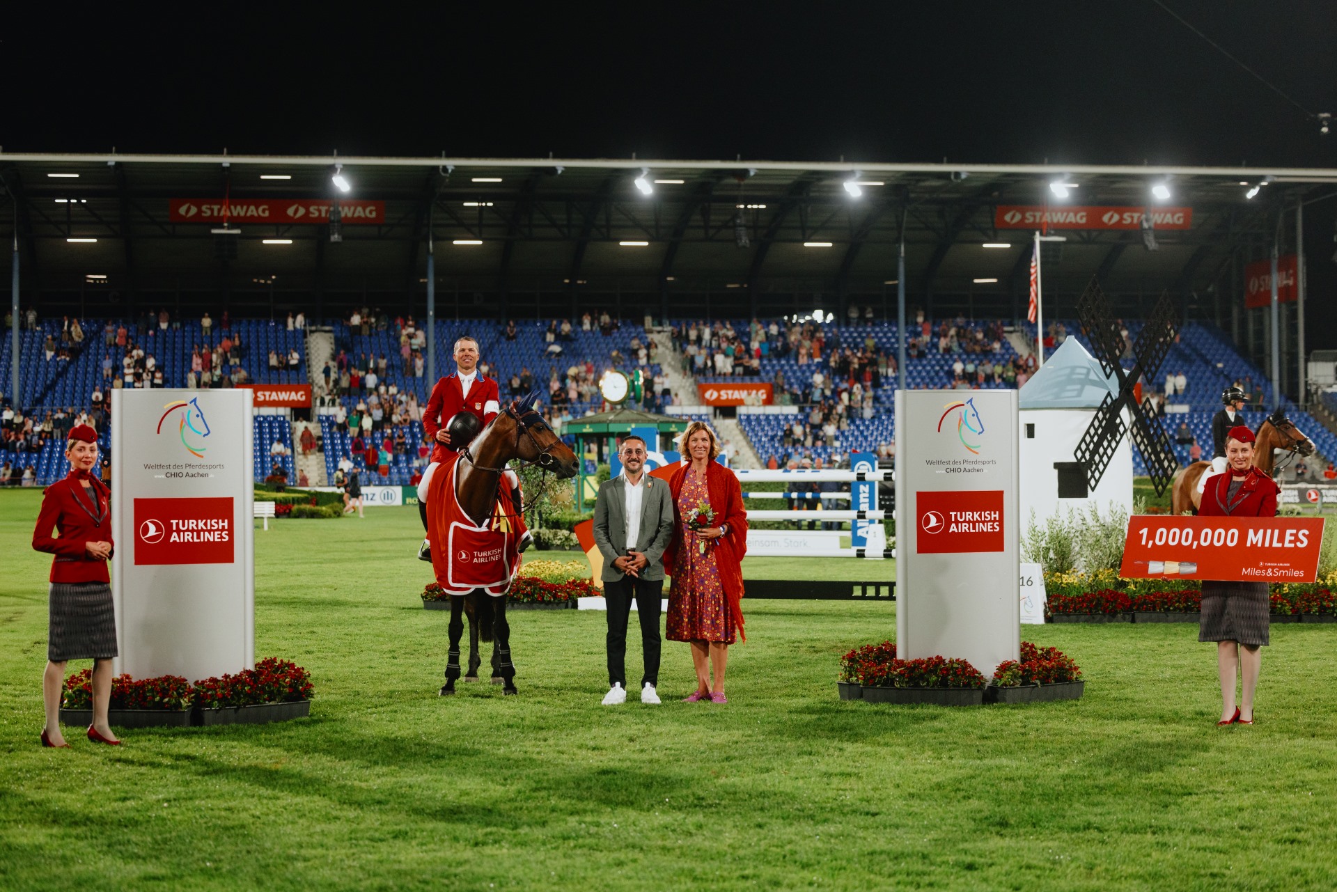Prize-giving ceremony in the show jumping arena at the CHIO Aachen at night. In the foreground are people and a horse with a winning blanket, flanked by two large boards with the Turkish Airlines logo. One person is holding a sign with the inscription ‘1,000,000 MILES’. The stands with spectators can be seen in the background.