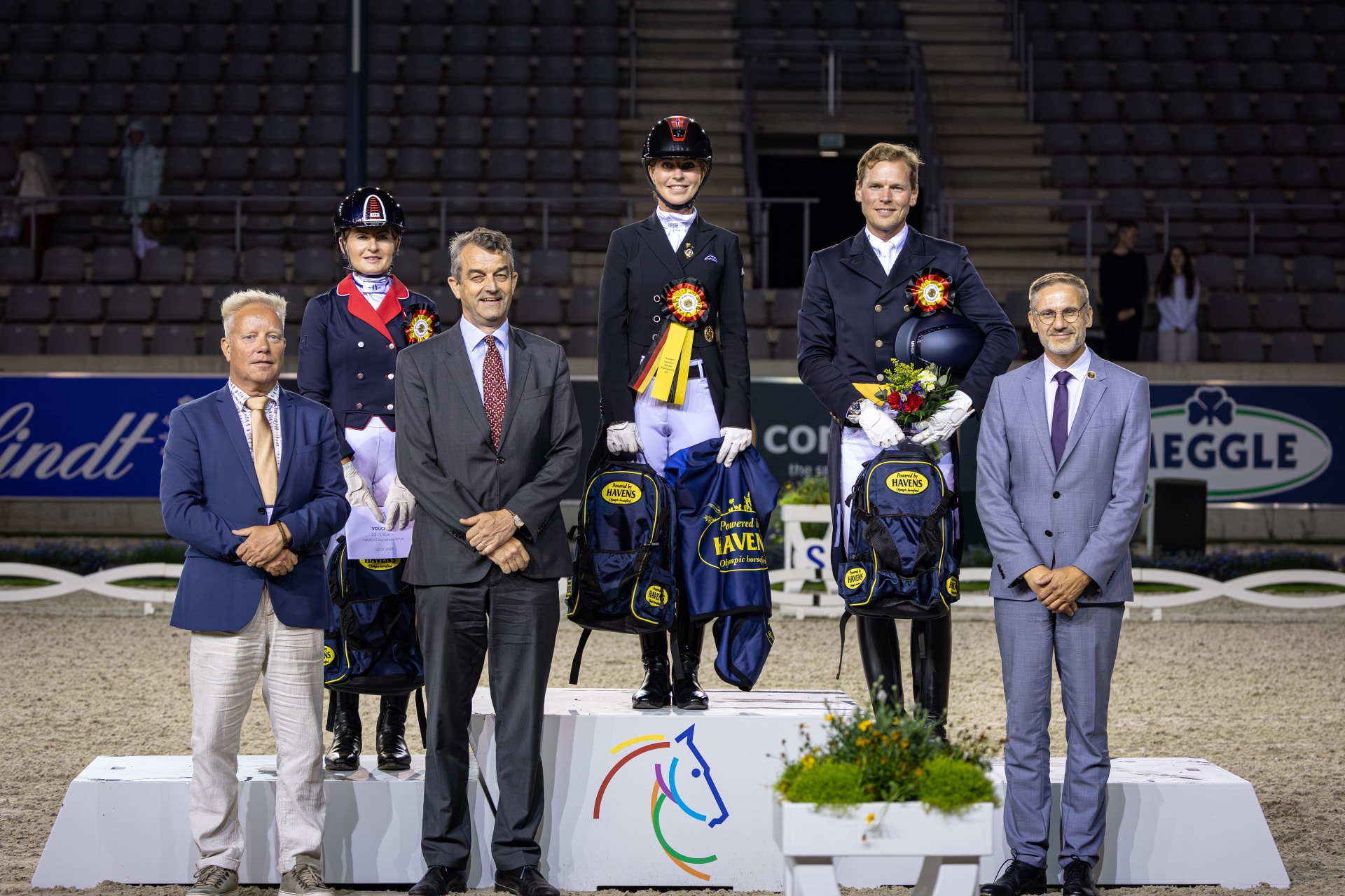 Prize-giving ceremony at an equestrian event in a stadium: Three riders in competition attire and helmets stand on the podium, surrounded by four officials in suits. The riders are wearing rosettes and holding bouquets and winner’s prizes. In the background, there are stands and advertising banners.