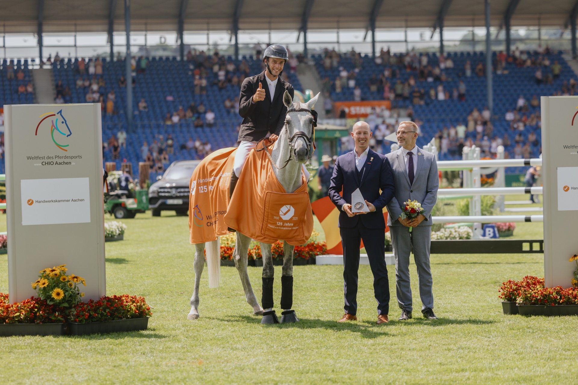 A rider sits on a white horse wearing an orange winner's blanket with the inscription ‘Handwerkskammer Aachen 125 Jahre’. Standing next to the horse are two elegantly dressed men, one holding a bouquet of flowers, the other a trophy. In the background are grandstands with spectators and two large signs with the CHIO Aachen and Handwerkskammer Aachen logos. The scene takes place on the grass of a large equestrian stadium.