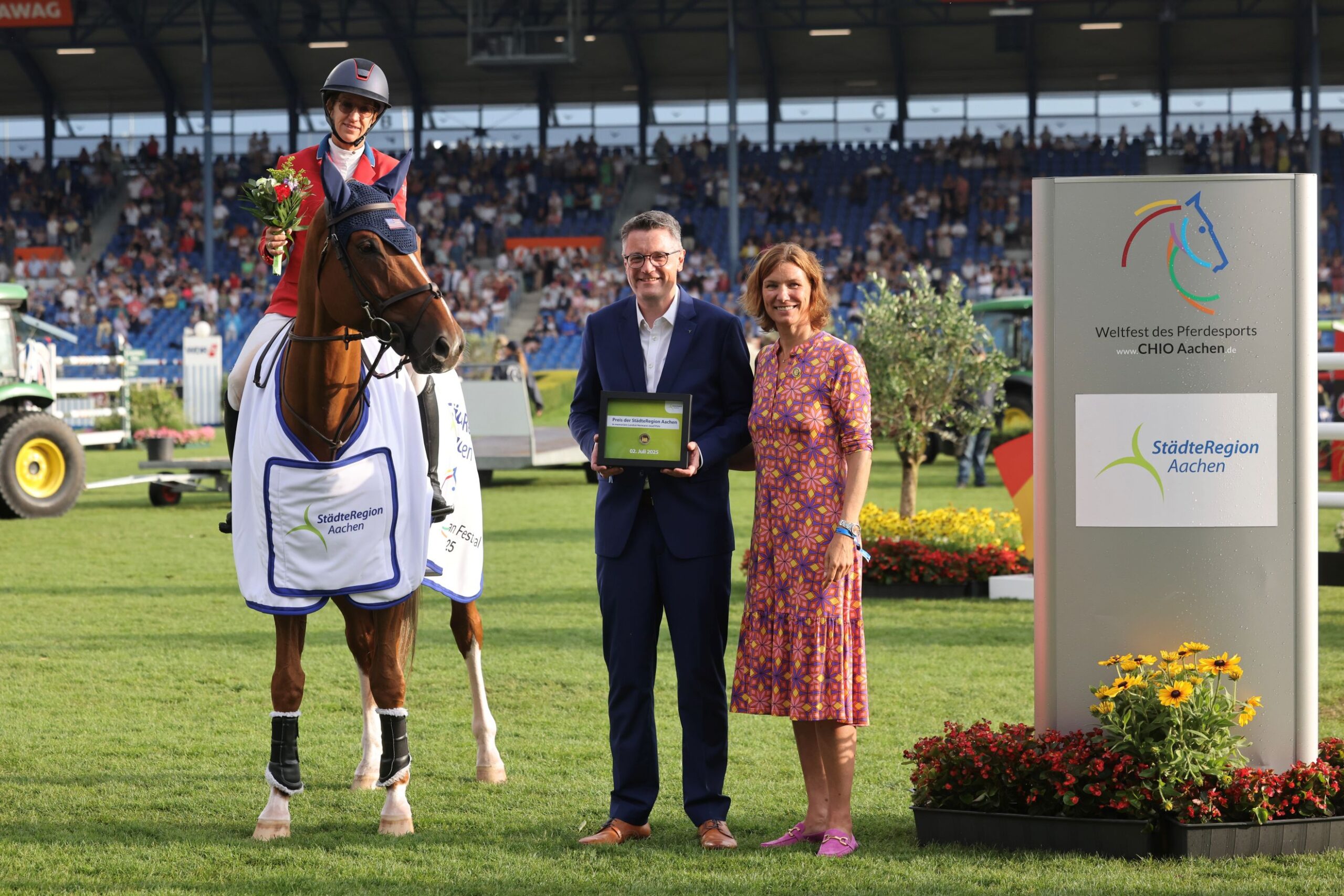A rider in a red jacket and helmet is sitting on a horse draped with a white winner's blanket labeled "StädteRegion Aachen" in a large stadium. The rider is holding a bouquet of flowers. Next to the horse, a man in a suit is holding a plaque, and a woman in a colorful dress stands beside him. In the background, there is a large sign with the CHIO Aachen logo and the words "Weltfest des Pferdesports" as well as "StädteRegion Aachen." The stadium stands are filled with spectators, and there are colorful flowers and greenery on the grass.