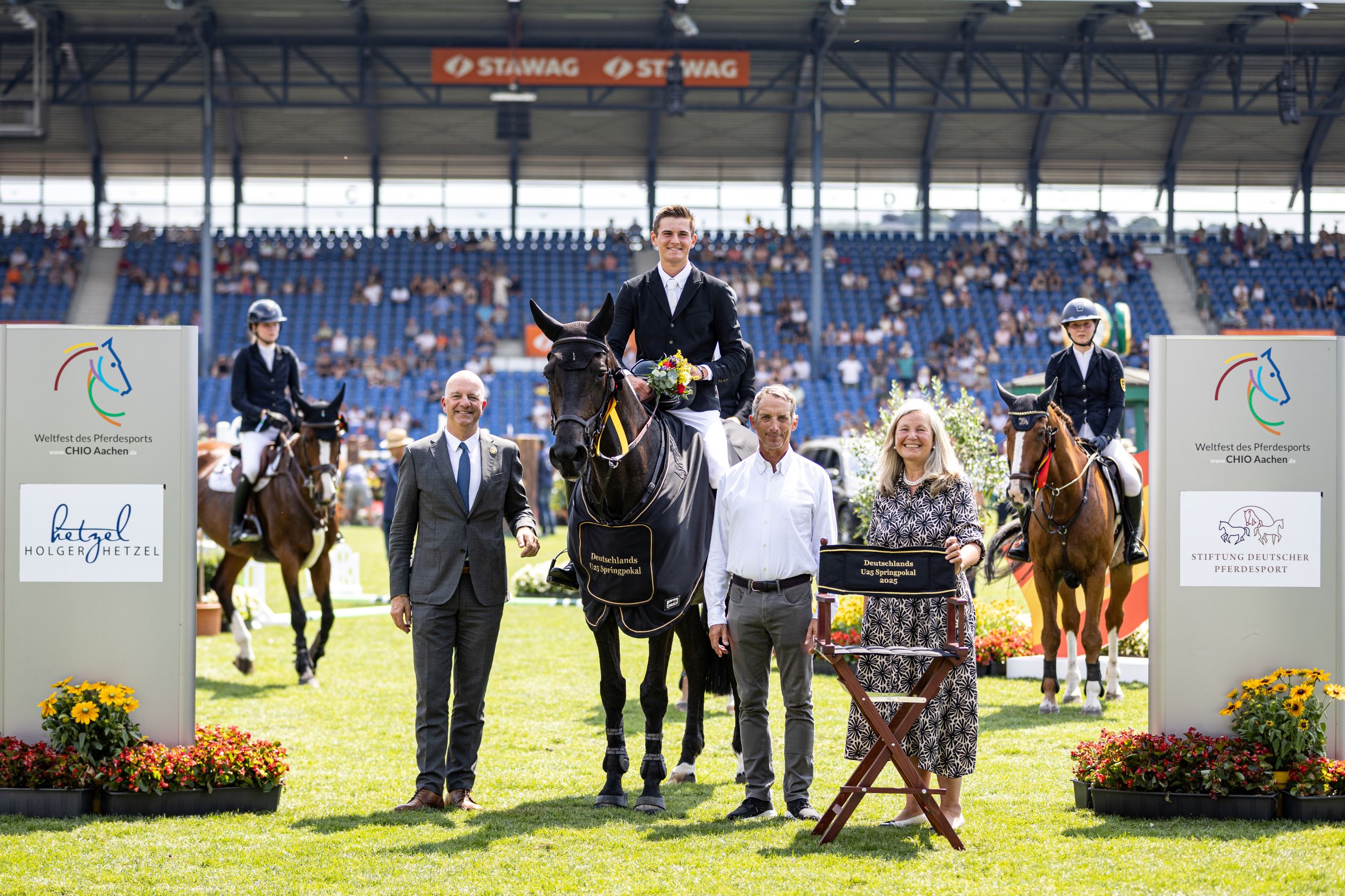 Winner’s photo at a show jumping event in the stadium: In the center, the winner in riding attire sits on a black horse, surrounded by three standing people. The horse wears a winner’s blanket with the inscription “Germany’s U25 Show Jumping Cup.” On the right, a woman holds a director’s chair with the same inscription. In the background, two more riders are on their horses. Large boards with CHIO Aachen logos and sponsors, decorated with flowers, are placed in the foreground on both sides. The stands in the background are filled with spectators.