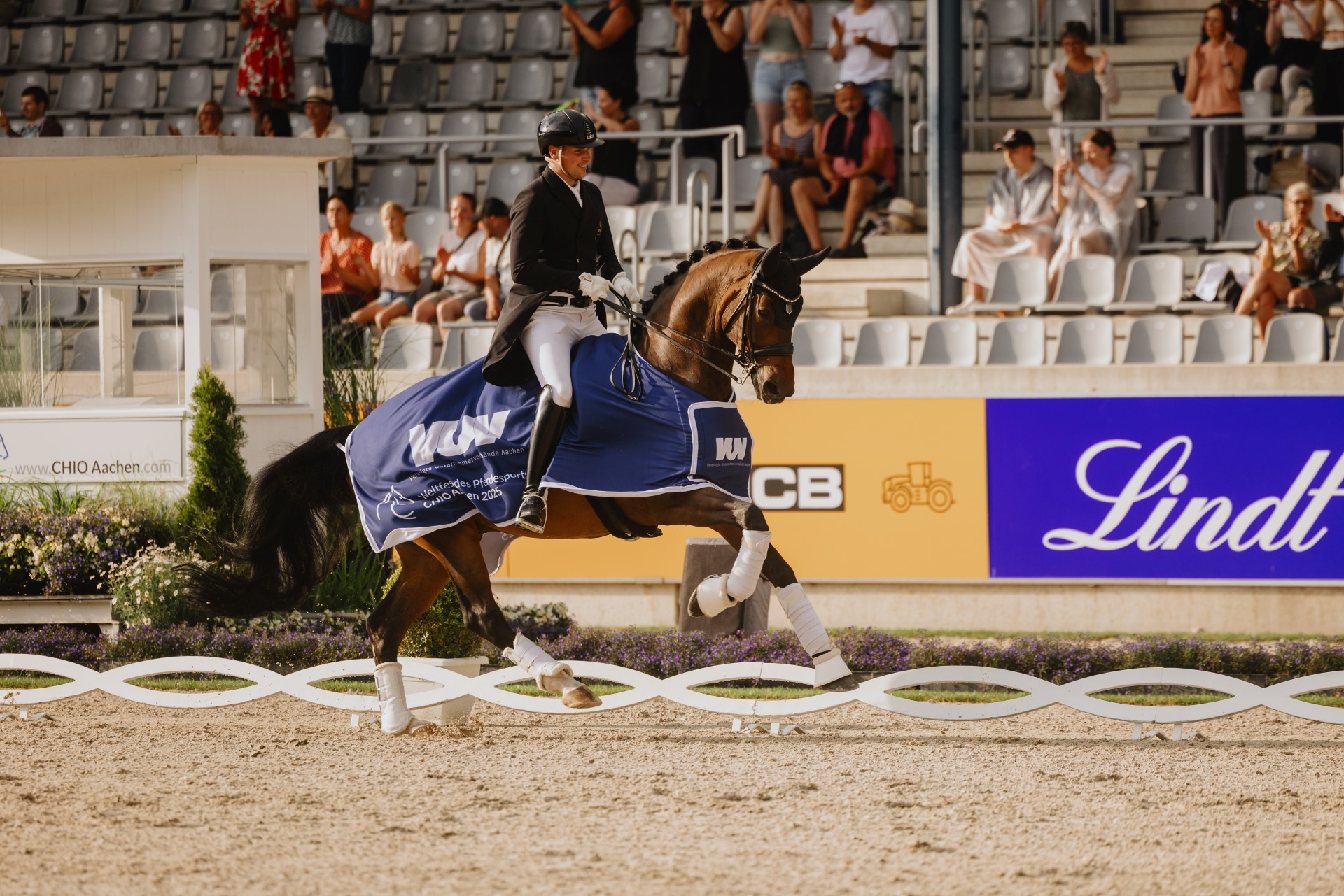 A rider in competition attire rides a dark brown horse wearing a winner's saddle pad with the inscription ‘World Equestrian Festival CHIO Aachen 2025’. In the background, spectators sit and stand in the stands, while advertising boards with sponsor adverts can be seen.