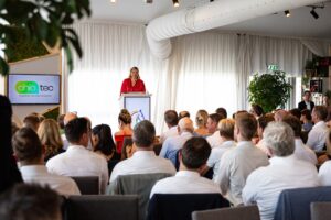 A woman in a red dress stands at a podium giving a speech to an audience seated in a modern, well-lit conference room. The audience consists of men and women dressed in business casual attire, facing the speaker. To the left, a screen displays the logo and slogan of “chio tec” with the tagline “together we can do better.” The room features white curtains, green plants, and a visible ventilation duct on the ceiling.