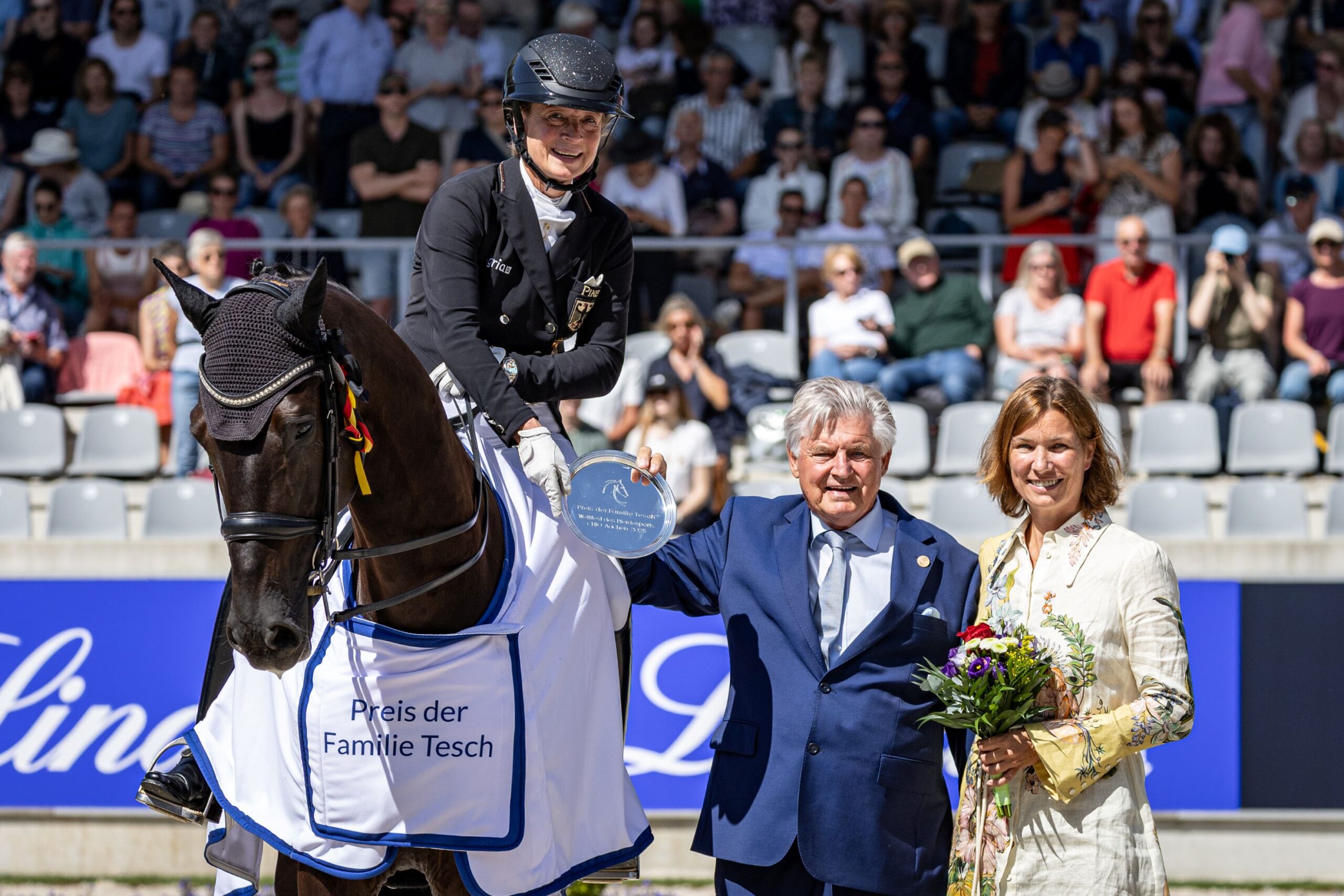 Eine Dressurreiterin reitet auf einem dunkelbraunen Pferd in einer Dressurprüfung. Im Hintergrund sind ein großes Anzeigeboard mit dem Namen „Isabell Werth“ und Zuschauer auf der Tribüne zu sehen. Die Reiterin trägt eine schwarze Jacke, weiße Reithose und einen Helm. Die Szenerie findet in einem Stadion mit Blumendekorationen und Dressurviereck statt.