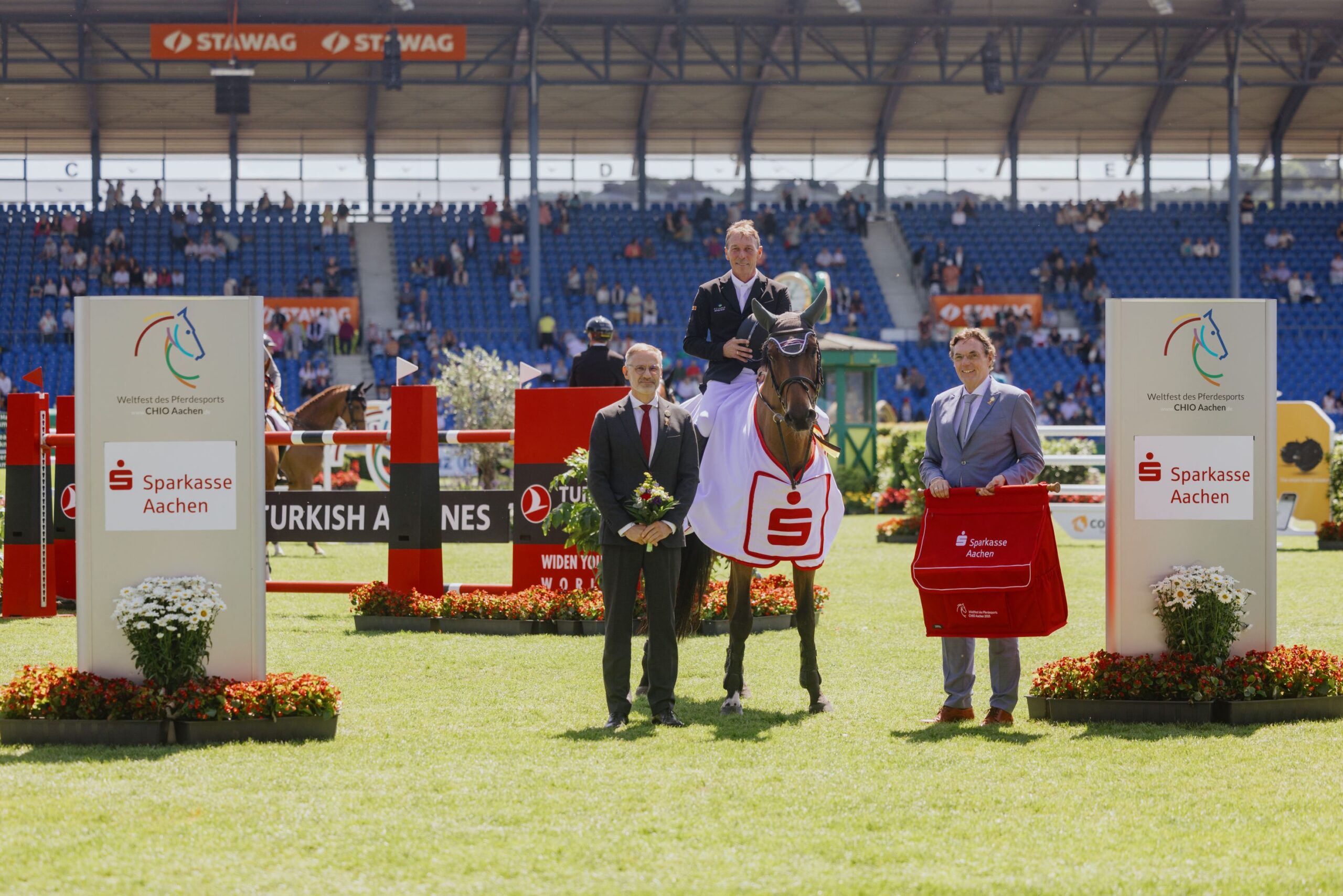 Siegerehrung im Sparkassen-Youngsters-Cup: Ein Reiter sitzt auf seinem Pferd, das eine Siegerdecke mit Sparkasse-Logo trägt. Zwei Personen stehen links und rechts daneben, eine hält Blumen, die andere eine rote Tasche. Im Hintergrund sind Hindernisse, Werbetafeln und Zuschauer auf den Tribünen zu sehen.