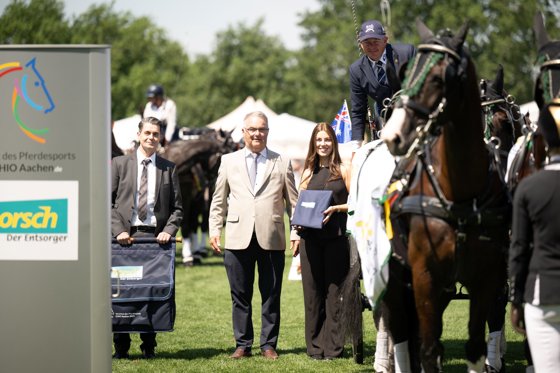 Mehrere Personen und ein Gespann mit Pferden stehen auf einem Turnierplatz beim CHIO Aachen. Links ist ein Schild mit dem Logo des CHIO Aachen und dem Sponsor Horsch zu sehen. Eine Frau hält eine blaue Mappe, ein Mann im Anzug hält eine Pferdedecke, und ein weiterer Mann steht in der Mitte. Im Hintergrund sind weitere Pferde und Teilnehmer zu erkennen.