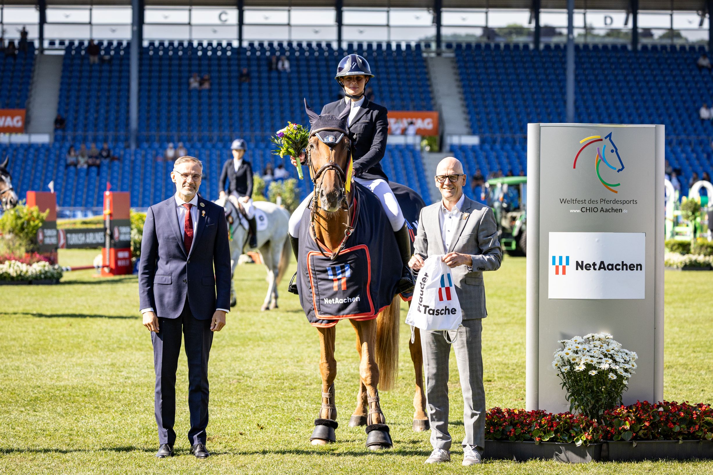 Drei Personen und ein Pferd stehen auf einem Turnierplatz im Stadion. Die Person in der Mitte sitzt auf einem braunen Pferd, das eine Siegerdecke mit dem Logo von NetAachen trägt. Links steht eine Person im Anzug, rechts eine Person im hellen Sakko mit einer NetAachen-Tasche. Im Hintergrund sind weitere Reiterinnen und Reiter, Zuschauer auf den Tribünen sowie ein Schild mit dem Logo des CHIO Aachen und NetAachen zu sehen.