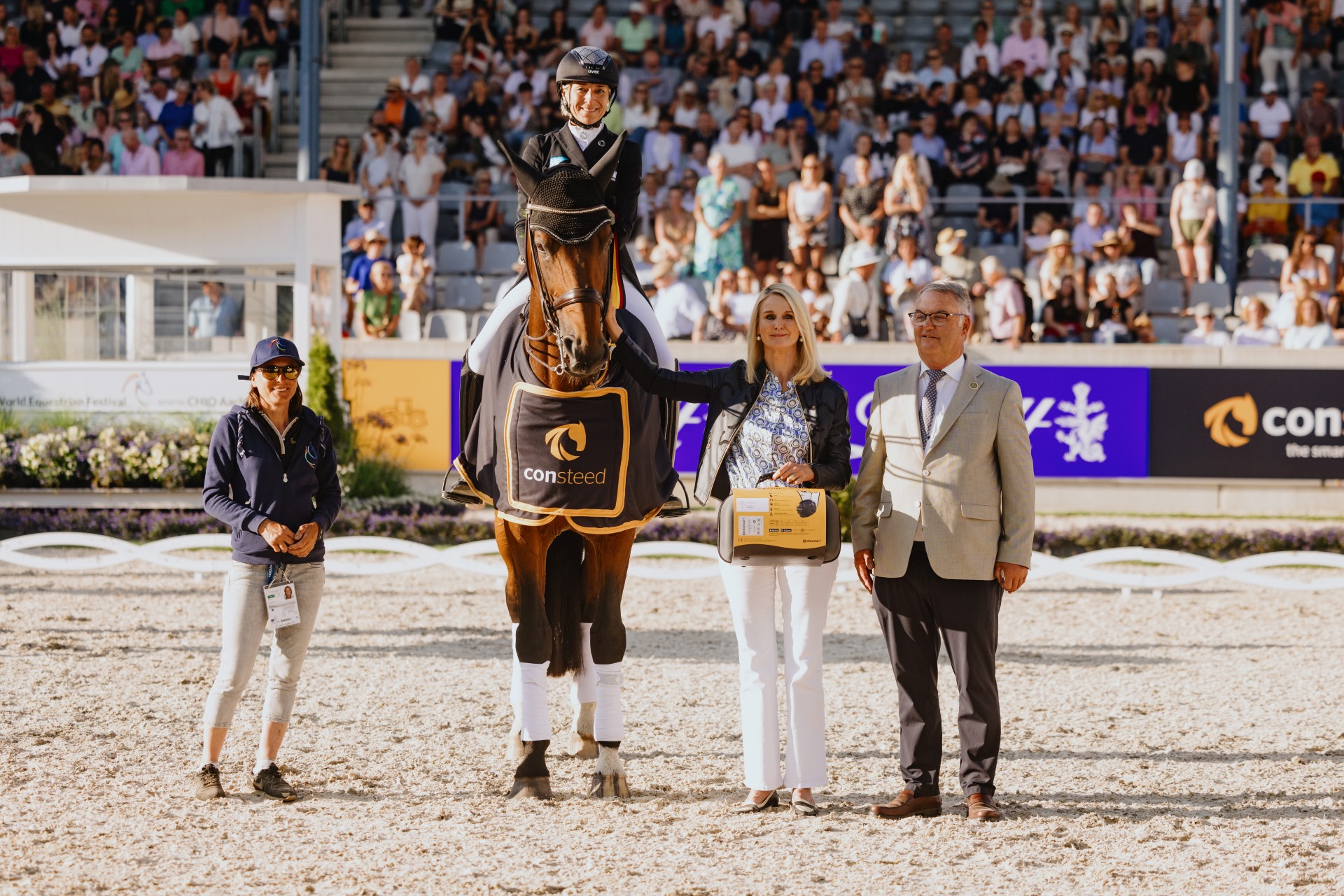Eine Siegerehrung bei einem Reitturnier: Eine Reiterin sitzt auf einem Pferd, das eine schwarze Siegerdecke mit dem Logo und Schriftzug „consteed“ trägt. Neben dem Pferd stehen drei Personen, von denen eine eine gelbe Geschenkbox hält. Im Hintergrund ist eine Tribüne mit zahlreichen Zuschauern zu sehen, die das Geschehen beobachten. Die Szene spielt sich auf einem sandigen Reitplatz ab.