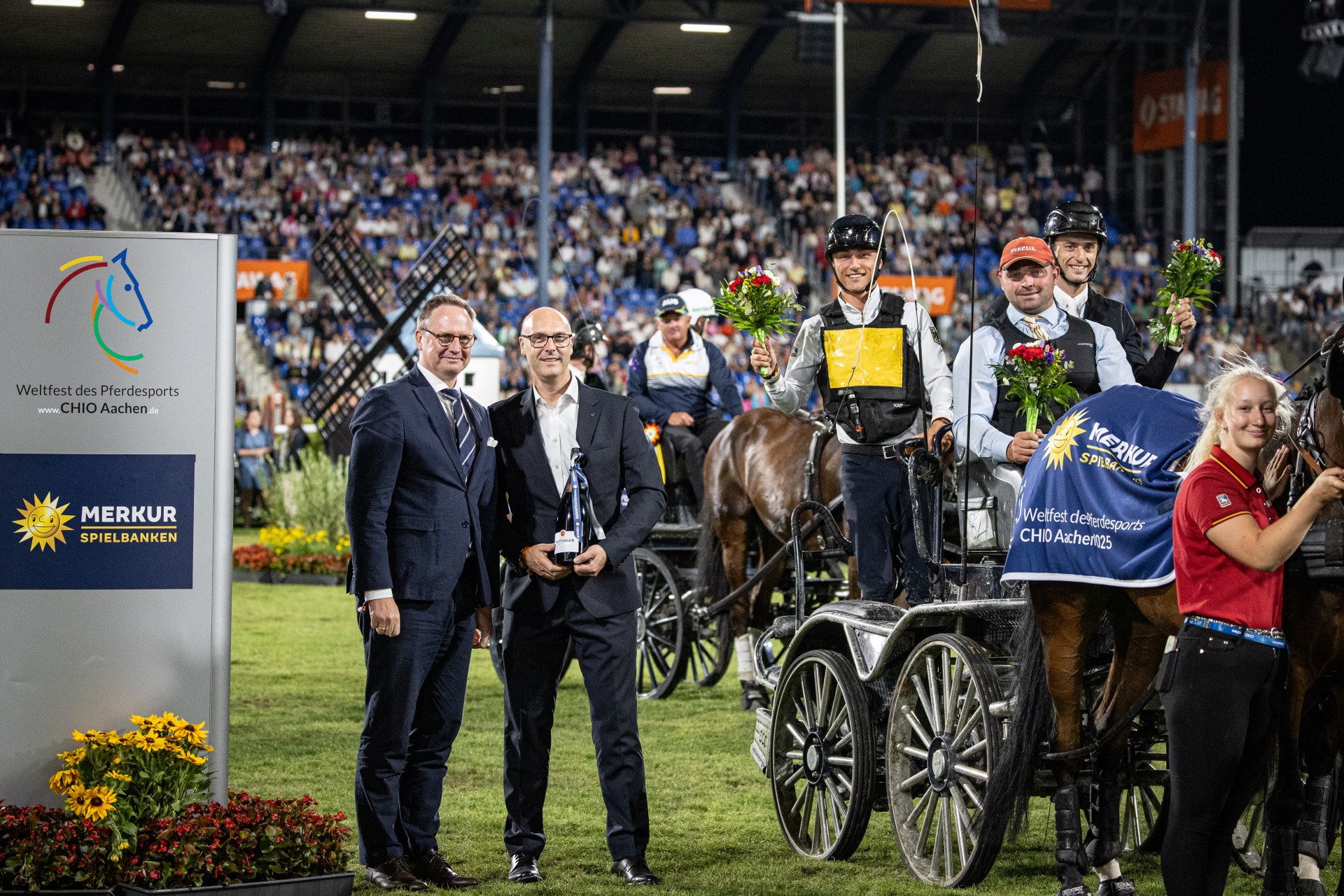 Several team drivers stand with their horses and carriages on the grass of a stadium. They are wearing winning blankets with the Merkur Spielbanken logo and are holding bouquets of flowers. On the left of the picture is a large board with the CHIO Aachen and Merkur Spielbanken logos. In the background, numerous spectators can be seen in the stands.