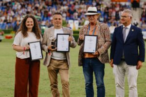 Four people stand on a grass pitch in front of a full grandstand. Three of them are holding framed certificates in their hands, one person is also holding a trophy. The people are festively dressed, one is wearing a white hat and a colorful jacket. In the background, many spectators can be seen in the stands.