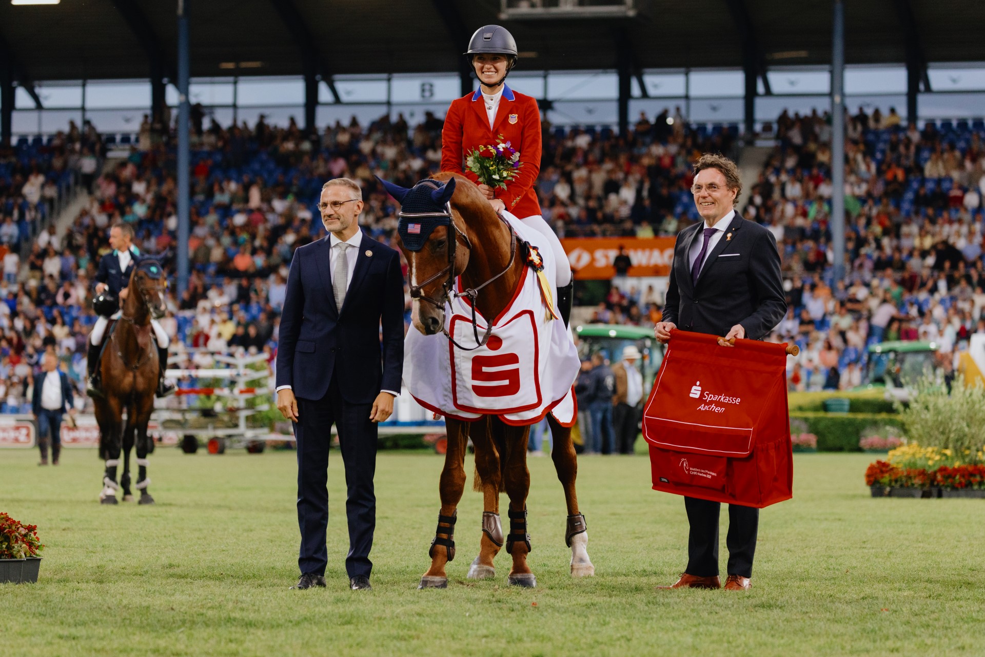 A rider in a red jacket sits on a horse wearing a white winner's saddle pad with a red Sparkasse logo. She is holding a bouquet of flowers. To the left and right of the horse are two men in suits, one of whom is holding a large red bag with the inscription “Sparkasse Aachen”. A full grandstand can be seen in the background, other horses and people are on the showground.
