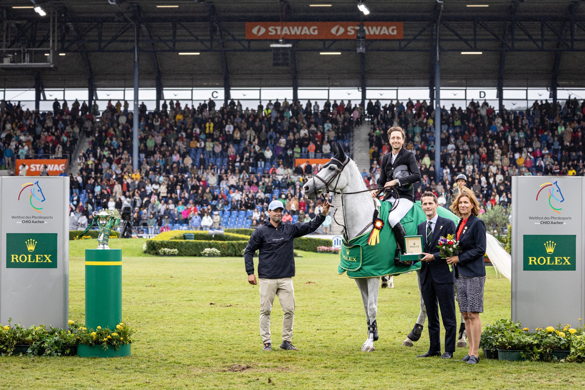 Bei einer Siegerehrung beim CHIO Aachen steht ein Reiter im Turnierkleidung auf einem Schimmel mit Siegerdecke und Schleife im großen Stadion. Neben dem Pferd stehen mehrere Personen, eine von ihnen hält eine Rolex-Uhr, eine andere einen Blumenstrauß. Im Hintergrund ist eine große Tribüne mit vielen Zuschauern zu sehen, links und rechts stehen grüne Rolex-Säulen mit der Trophäe und dem Logo des CHIO Aachen.