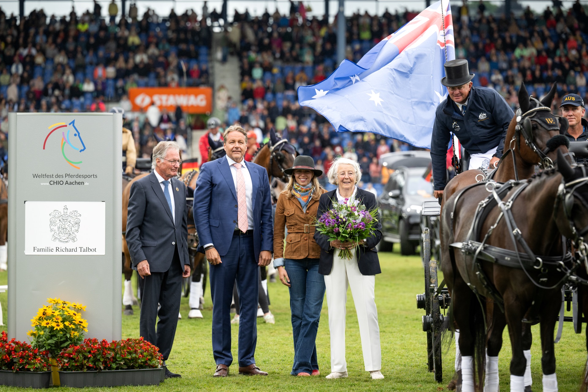 Siegerehrung beim CHIO Aachen: Im Vordergrund stehen mehrere Personen auf dem Rasen des Hauptstadions, darunter eine ältere Dame mit einem Blumenstrauß. Rechts im Bild ein Kutscher mit Zylinder auf einem Pferdegespann, der eine australische Flagge trägt. Links ist ein Schild mit dem Logo des CHIO Aachen und dem Namen 'Familie Richard Talbot' zu sehen. Im Hintergrund sind die voll besetzten Tribünen des Stadions zu erkennen