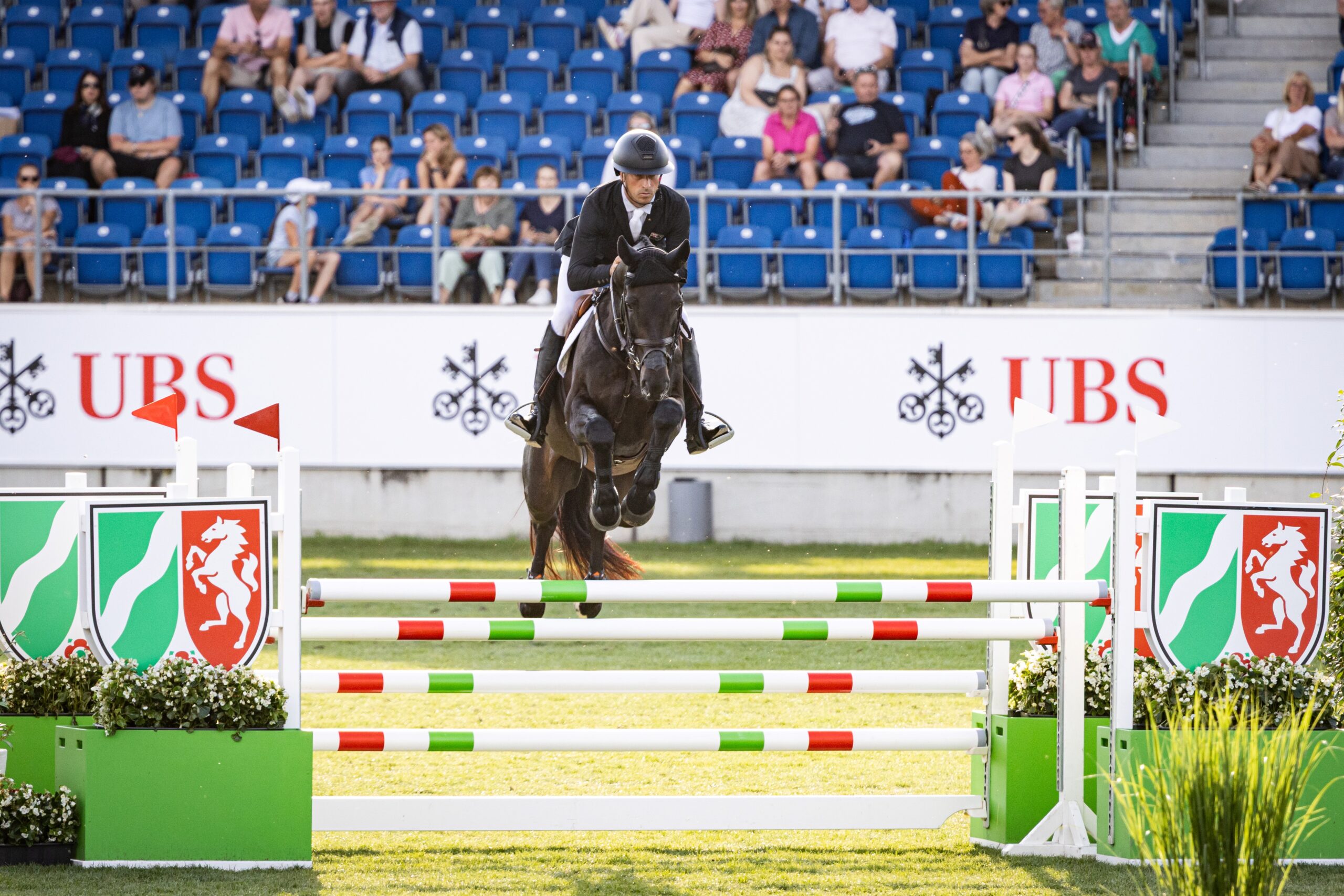 Ein Reiter springt mit seinem Pferd über ein farbiges Hindernis bei einem Springreitturnier im Stadion. Im Hintergrund sitzen Zuschauer auf blauen Tribünenplätzen, und Werbebanner mit UBS-Logos sind sichtbar.