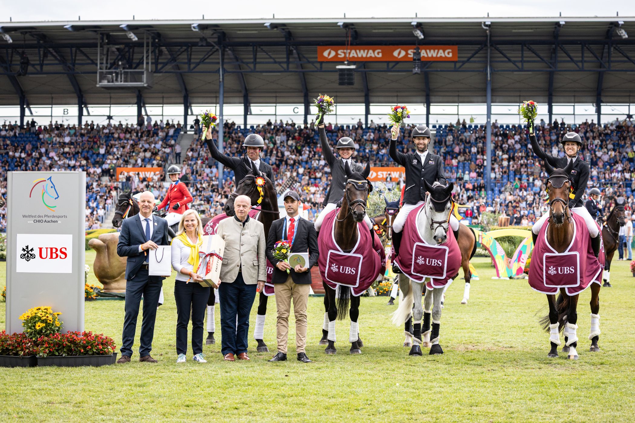 Award ceremony at the CHIO Aachen on the grass of the main stadium. Four riders sit on their horses in competition dress, wearing winning blankets with the UBS logo and holding up bouquets of flowers. Several people, including officials and guests of honor, stand in front of the horses with gifts and flowers. In the background, full grandstands with numerous spectators can be seen, on the left is a stele with the CHIO Aachen and UBS logo.