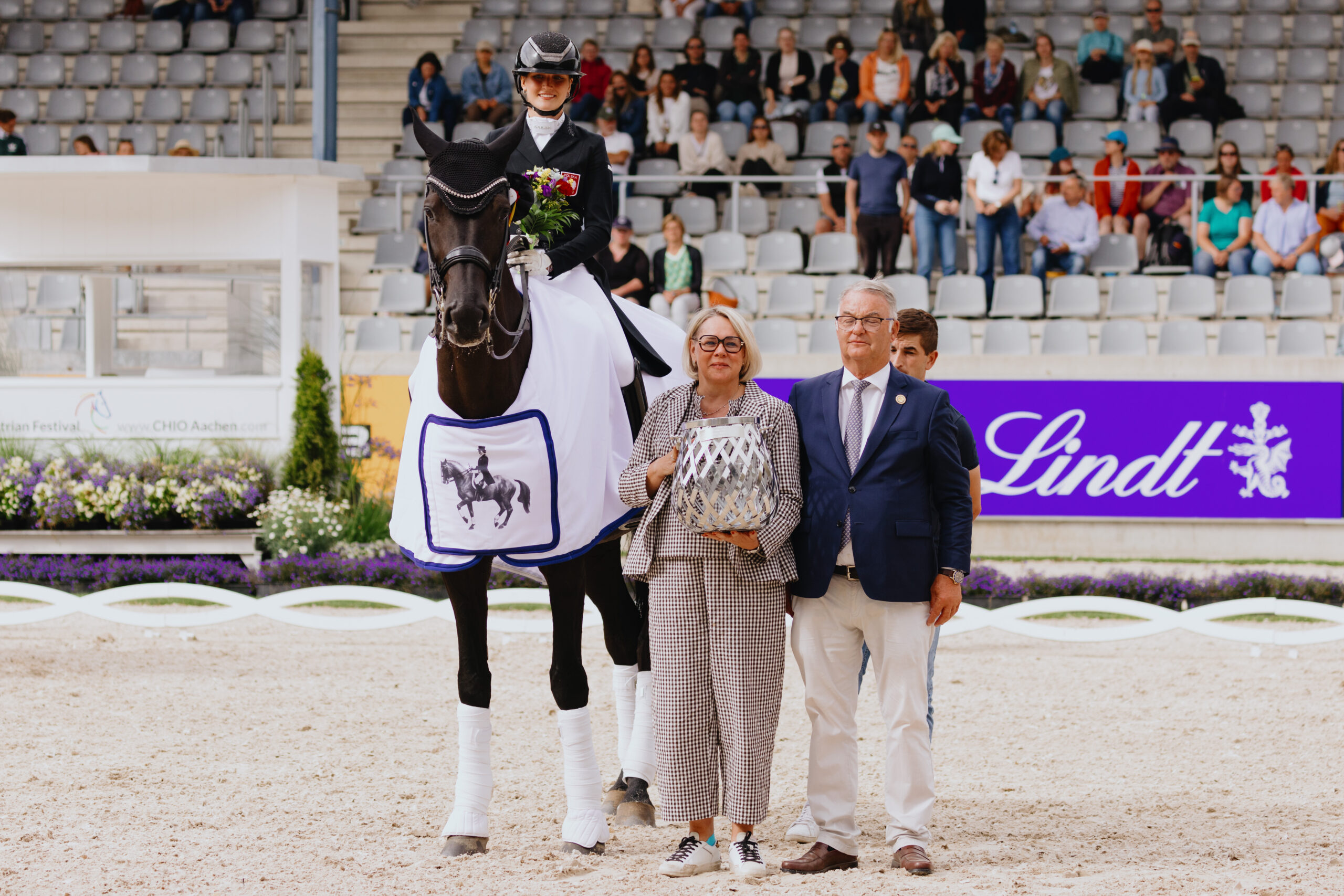 A rider is sitting on a dark brown horse, which is wearing a white blanket and bandages. Two people are standing next to the horse on sandy ground, one of them holding a large silver trophy. Spectators on stands and a purple Lindt advertising banner can be seen in the background. The scene takes place in an equestrian stadium, decorated with flowers at the edge of the arena.