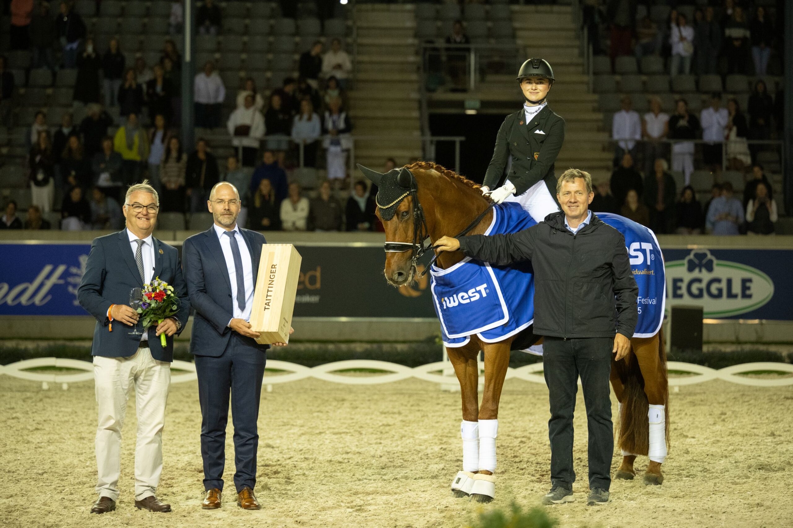 A rider sits on a brown horse with a blue winner's blanket in an illuminated riding arena. Standing next to the horse are three men, one holding a bouquet of flowers, another a large wooden box labelled ‘Taittinger’. Spectators in the stands can be seen in the background.