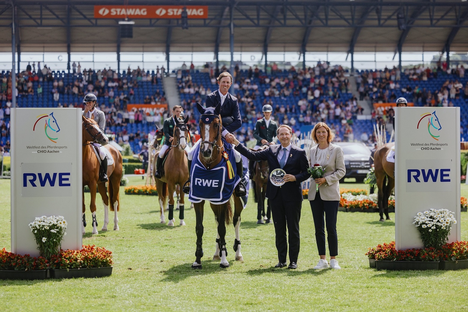 Siegerehrung bei einem Reitturnier im Stadion: Ein Reiter sitzt auf einem Pferd mit einer blauen Siegerdecke mit RWE-Logo. Zwei Personen stehen daneben, eine hält einen Glasteller, die andere einen Blumenstrauß. Im Hintergrund stehen weitere Reiterinnen und Reiter auf ihren Pferden sowie Zuschauer auf den Tribünen. Links und rechts sind große Tafeln mit dem CHIO Aachen-Logo und dem Schriftzug RWE zu sehen.