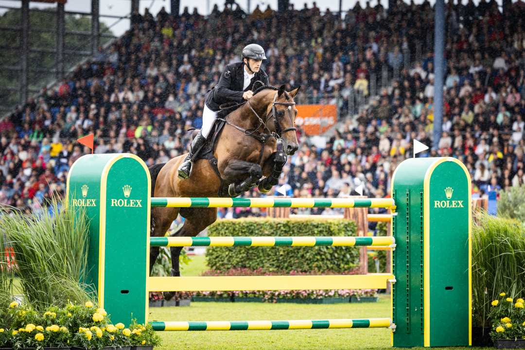A rider is jumping with his horse over a green and yellow obstacle at a show jumping competition. The Rolex logo is visible on the jumps. In the background, there is a large, crowded grandstand with spectators. It is raining lightly during the competition.