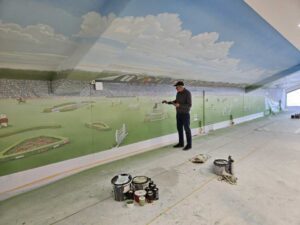 In a long interior space, an artist is painting a large-scale mural depicting a stadium and equestrian scene under a blue sky. Paint buckets and brushes lie on the floor, and light streams into the room through a window on the right.