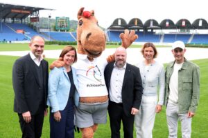 Gruppe von vier Frauen und Männern steht auf dem Rasen eines großen Reitstadions neben einem Pferdemaskottchen in braunem Fellkostüm. Das Maskottchen trägt ein weißes T Shirt mit buntem Pferdelogo und der Aufschrift „#chioaachen“ und breitet einen Arm winkend aus. Im Hintergrund sind leere blaue Tribünen, ein Springplatz und mehrere Nationalflaggen zu sehen.