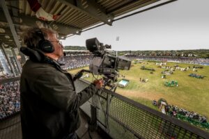 Kameramann mit großen Kopfhörern filmt mit einer professionellen TV-Kamera von einer überdachten Tribüne aus ein gut besuchtes Reitstadion; unten auf dem weitläufigen Rasenplatz sind Hindernisse und Parcours-Elemente aufgebaut, rundherum sitzen zahlreiche Zuschauer auf den Rängen.