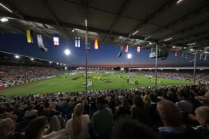 Panoramablick in ein voll besetztes Reitstadion bei Abenddämmerung: Unter dem überdachten Tribünendach mit zahlreichen Länderflaggen blicken tausende Zuschauer auf den hell ausgeleuchteten Rasenplatz mit mehreren Springhindernissen.