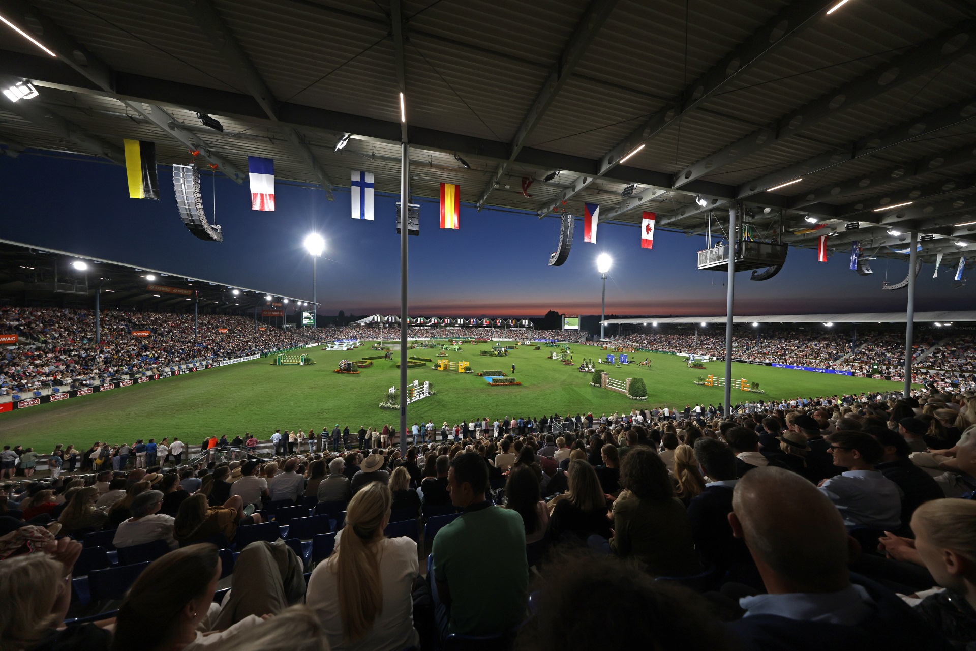 Panoramablick in ein voll besetztes Reitstadion bei Abenddämmerung: Unter dem überdachten Tribünendach mit zahlreichen Länderflaggen blicken tausende Zuschauer auf den hell ausgeleuchteten Rasenplatz mit mehreren Springhindernissen.