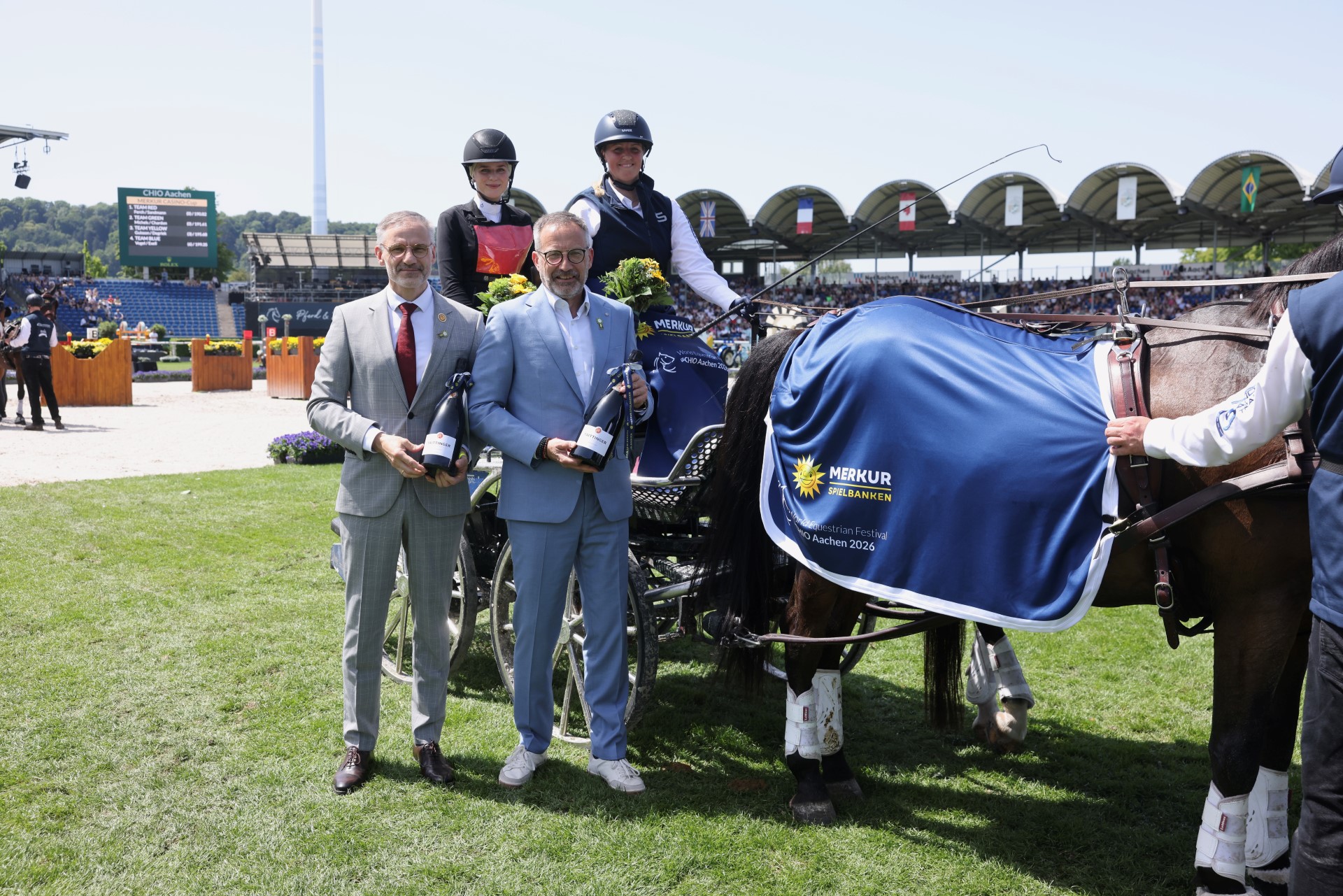 Siegerehrung beim Pferdesportturnier im Stadion: Zwei Herren im Anzug halten Sektflaschen, dahinter sitzen zwei Fahrerinnen in Kutschen-Outfits auf einem Gespann. Rechts im Bild steht ein Pferd mit blauer Siegerdecke mit dem Logo „MERKUR SPIELBANKEN“ und der Aufschrift „World Equestrian Festival CHIO Aachen 2026“. Im Hintergrund sind Tribünen mit Zuschauern, Flaggen und ein Anzeigeboard zu sehen.