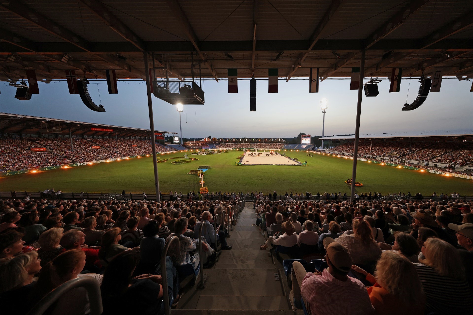 Weitwinkelaufnahme eines abendlichen Reitstadions, dicht gefüllte Tribünen im Vordergrund, im hell ausgeleuchteten Rasenoval in der Mitte eine sandfarbene Dressurfläche mit mehreren Pferden und Reitern, umgeben von Flutlichtmasten und Länderflaggen unter dem Stadiondach.