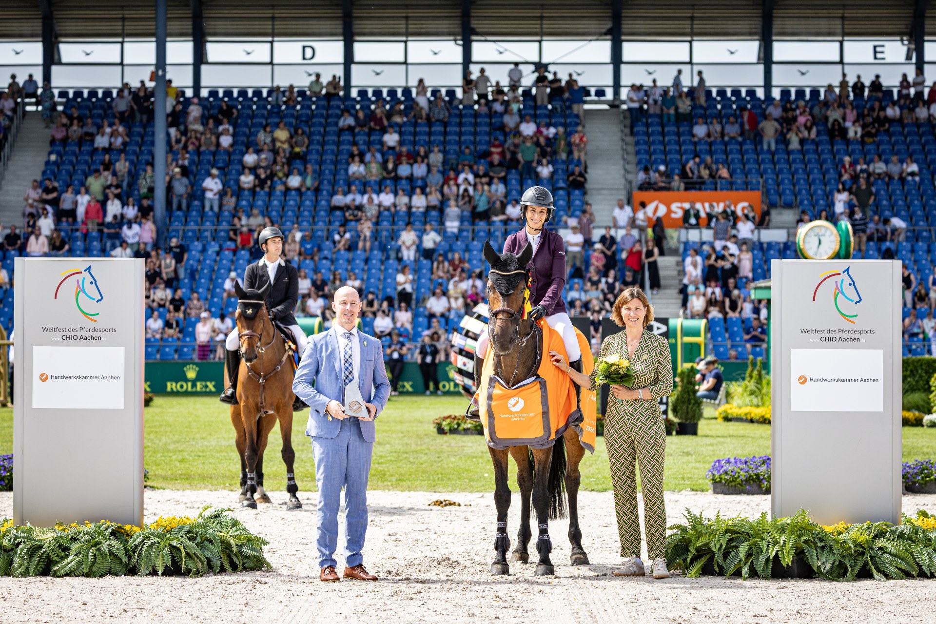 Siegerehrung beim CHIO Aachen in einem Springreitstadion: Eine Reiterin auf einem braunen Pferd mit orangefarbener Siegerdecke der Handwerkskammer Aachen steht zwischen zwei grauen Tafeln mit dem Logo „Weltfest des Pferdesports CHIO Aachen“. Links sitzt ein weiterer Reiter auf seinem Pferd, vorne stehen zwei Offizielle mit Pokal und Blumen. Im Hintergrund sind die Tribünen mit vielen Zuschauern zu sehen.